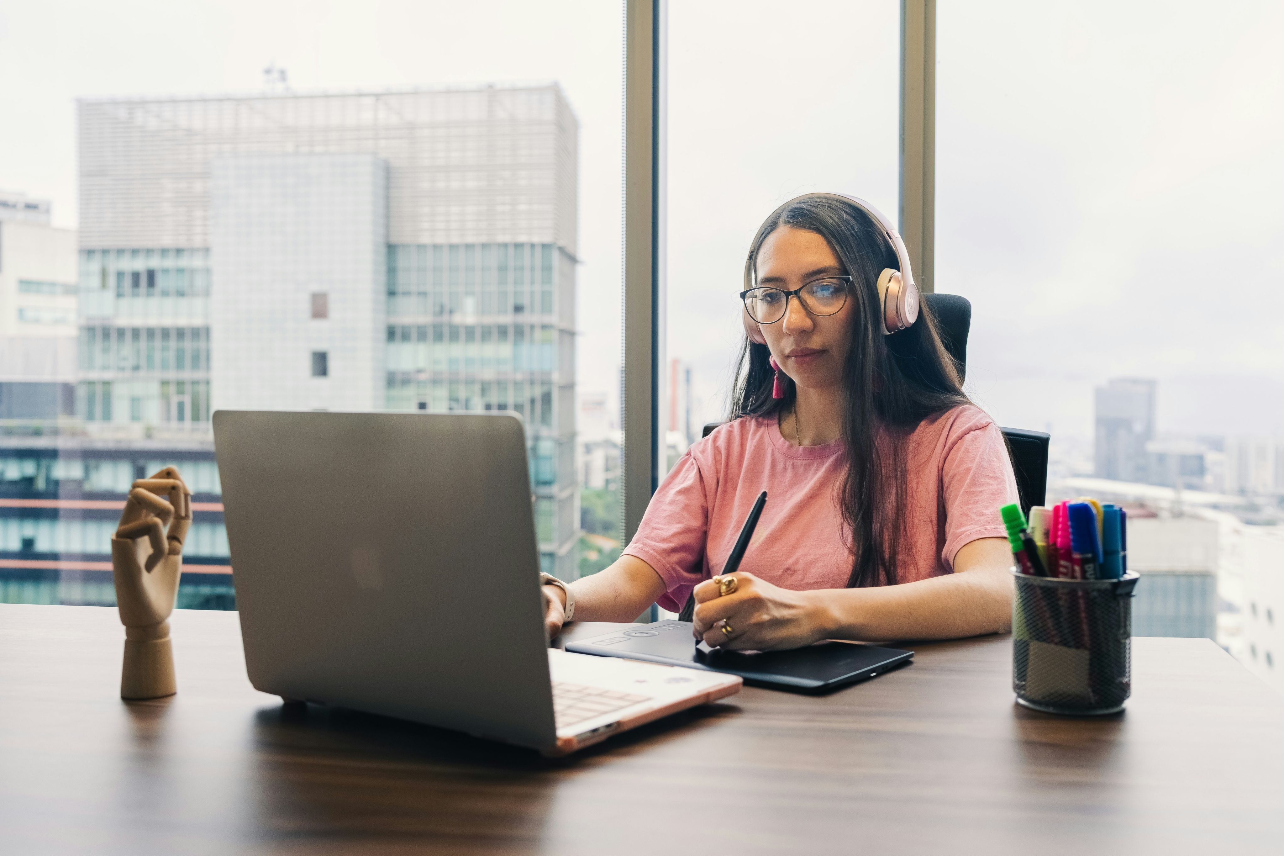 Woman working on laptop with headphones and stylus.